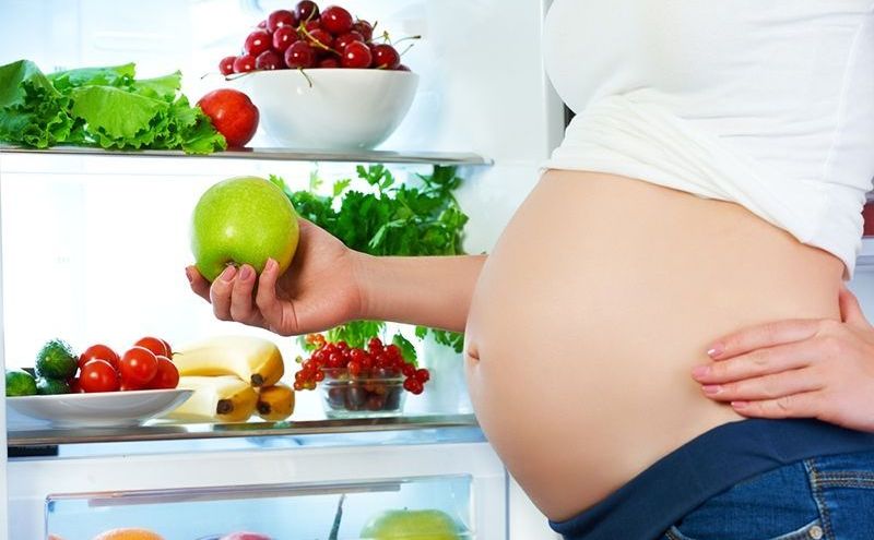 Schwangere Frau, die nahe Kühlschrank mit Obst und Gemüse steht