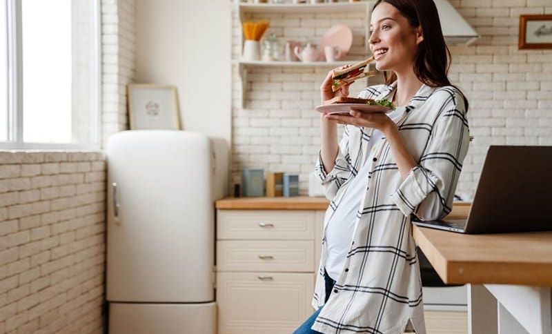 glückliche schwangere Frau isst ein Sandwich in der Küche