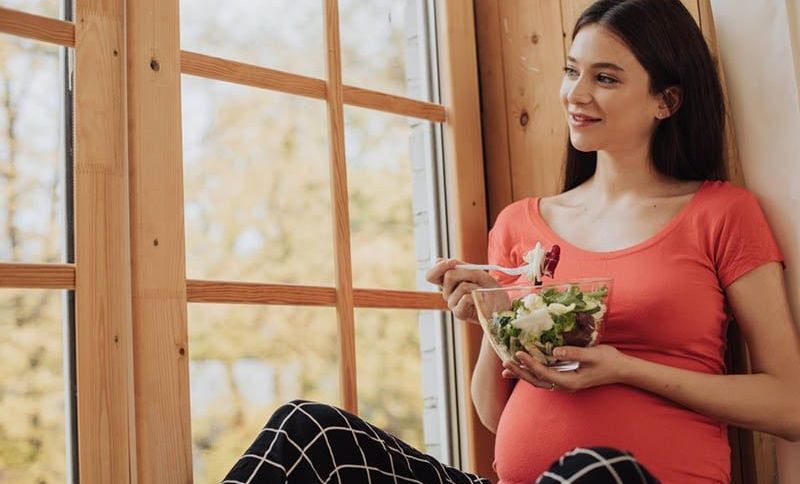 junge schwangere Frau sitzt am Fenster und isst einen Salat
