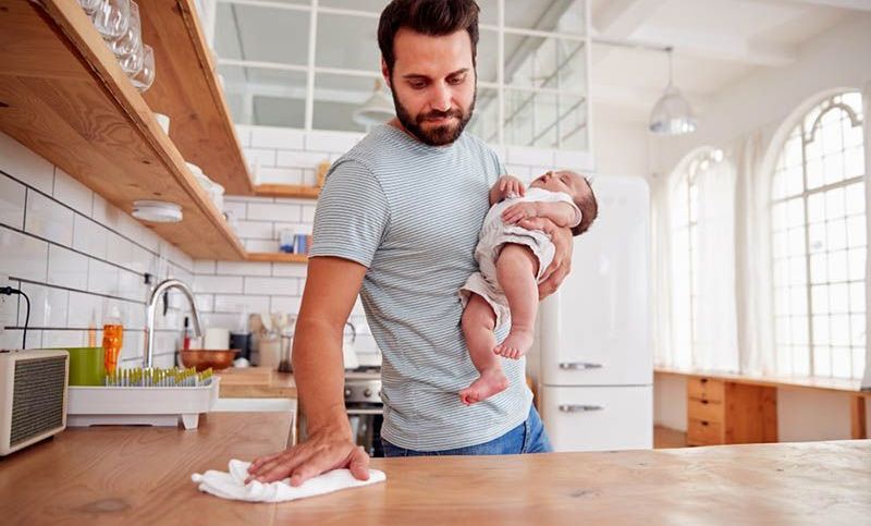 junger Vater hält schlafendes Baby und reinigt die Küchenoberfläche