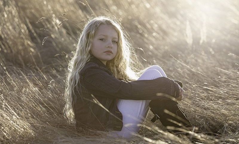 kleines Mädchen mit langen blonden Haaren, das auf dem Feld sitzt
